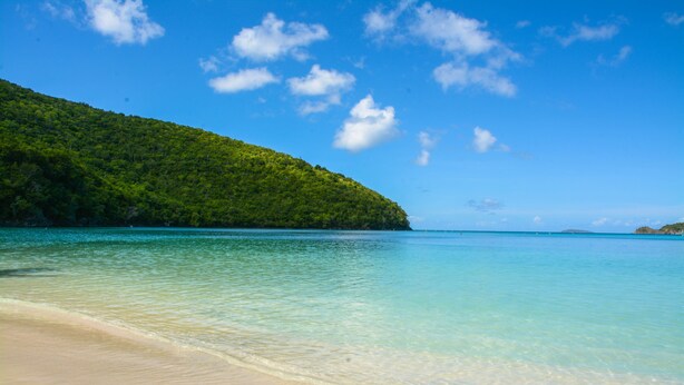 Une plage à l’eau turquoise et limpide, avec une colline verdoyante en arrière-plan.