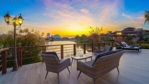 Une terrasse dotée de meubles en osier et d’une table, offrant une vue sur un plan d’eau et le panorama de la ville au coucher du soleil.