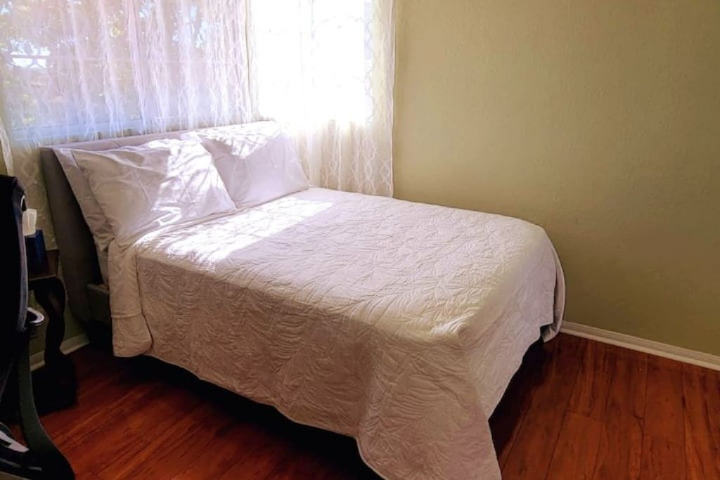 Una cama individual con colcha blanca en una habitación con suelo de madera y paredes verde claro.