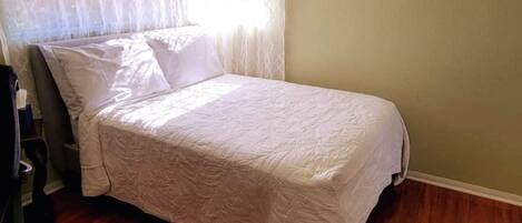 Una cama individual con colcha blanca en una habitación con suelo de madera y paredes verde claro.