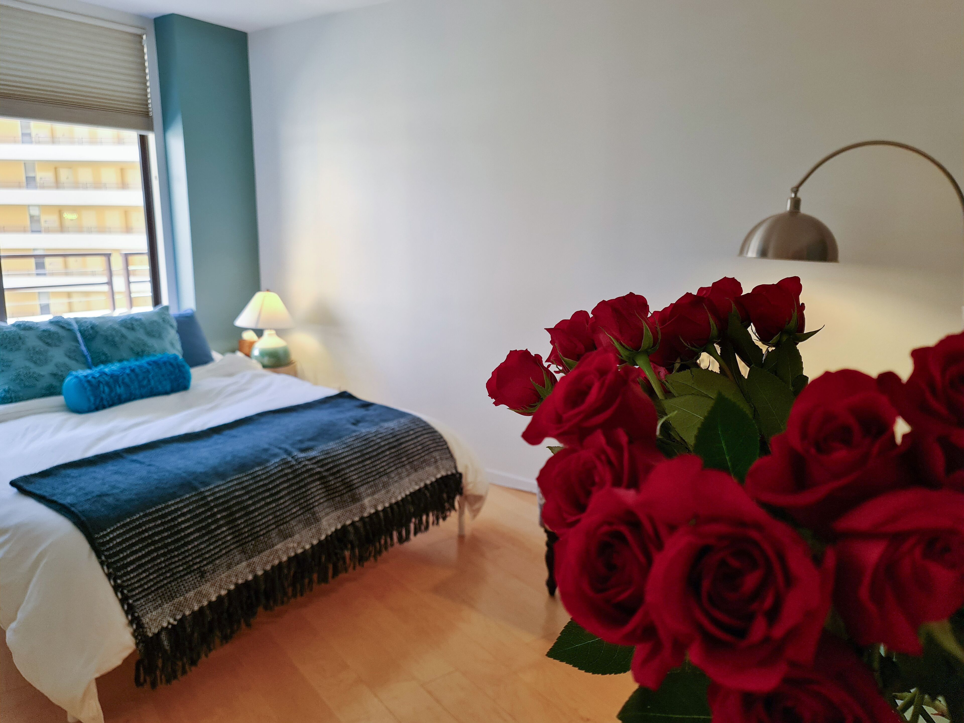 Un dormitorio con una cama, una lámpara de noche, una ventana con persianas y un jarrón de rosas rojas.