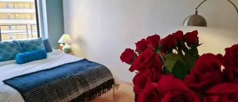 Un dormitorio con una cama, una lámpara de noche, una ventana con persianas y un jarrón de rosas rojas.