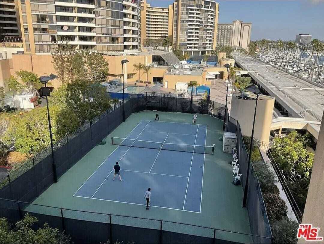 Una cancha de tenis al aire libre rodeada de edificios, con un estacionamiento visible al fondo.