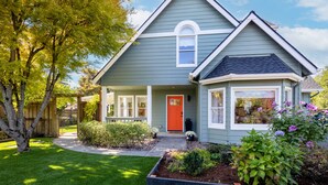 Une maison de plain-pied avec une porte rouge, un porche à l’avant et un petit jardin où poussent différentes plantes et fleurs.