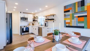 Une cuisine moderne avec des meubles blancs, un four à micro-ondes et un lave-vaisselle. On y trouve une table à manger dressée avec de l’assiette et des bols blancs, ainsi qu’une plante en pot posée sur la table.