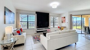 A modern living room with a white sofa, a glass coffee table, a flat-screen TV, and a large window with blinds. - Large 4-Bedroom 4-Bathroom Pool near attractions and Theme Parks (Kissimmee)
