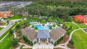 An aerial view of a residential area with a central swimming pool surrounded by houses, a well-maintained lawn, and a distant view of a cityscape. - Large 4-Bedroom 4-Bathroom Pool near attractions and Theme Parks (Kissimmee)