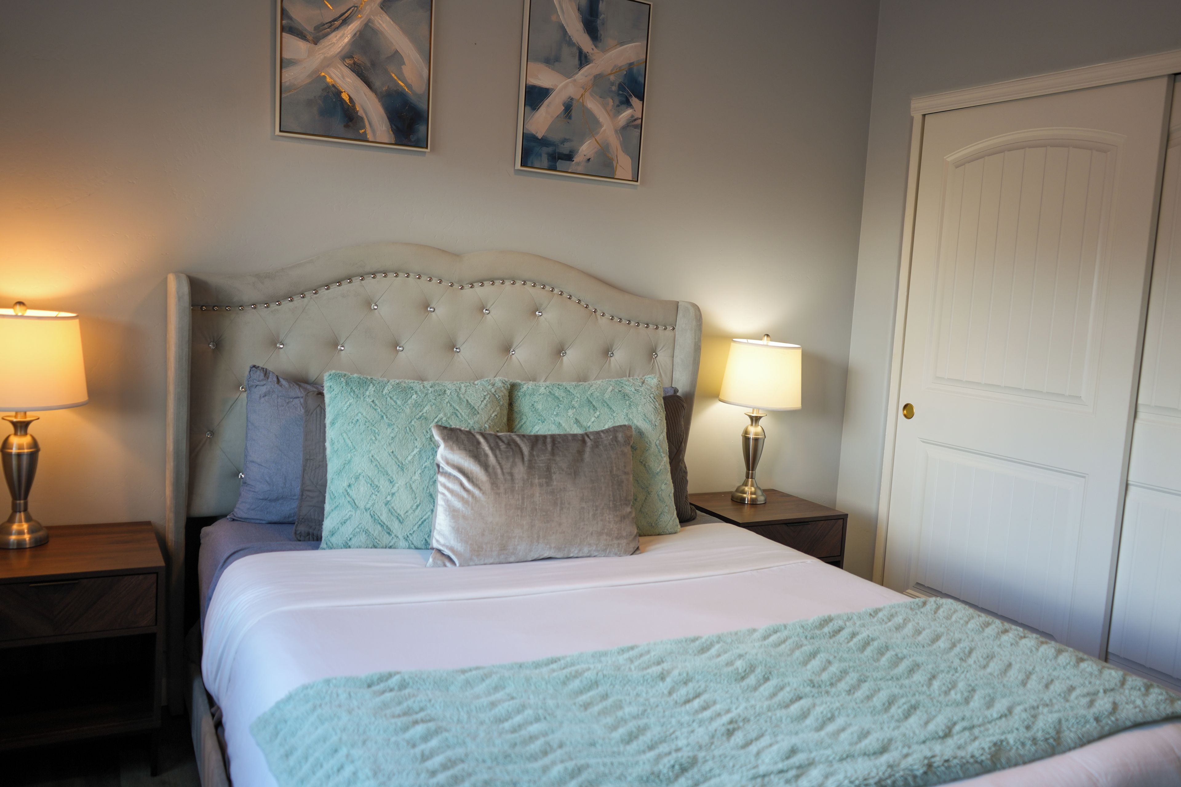 A bedroom with a tufted headboard, two bedside tables with lamps, a door, and two abstract paintings on the wall.