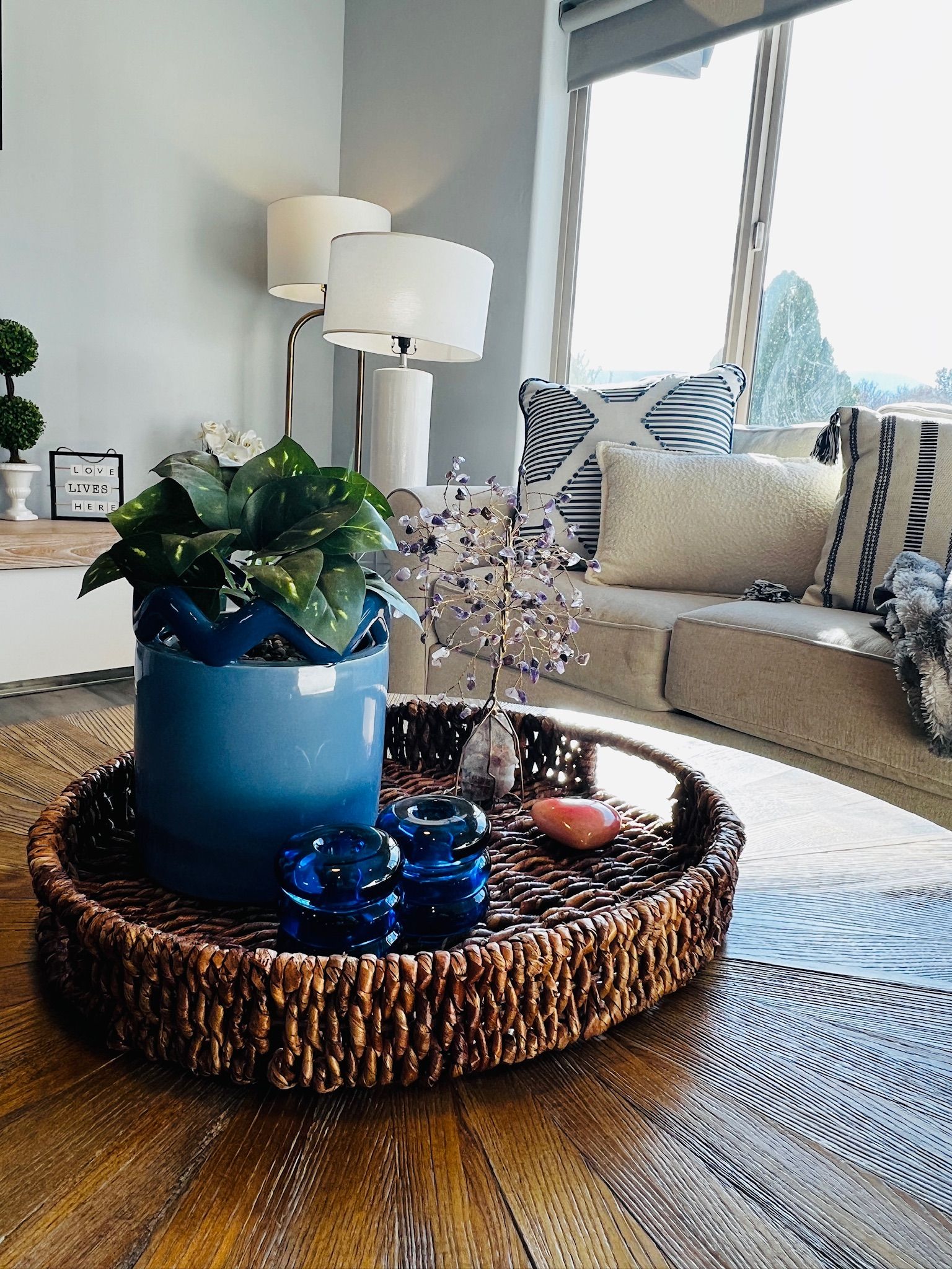 A living room with a blue potted plant, a woven tray, decorative jars, and a sofa with patterned cushions.