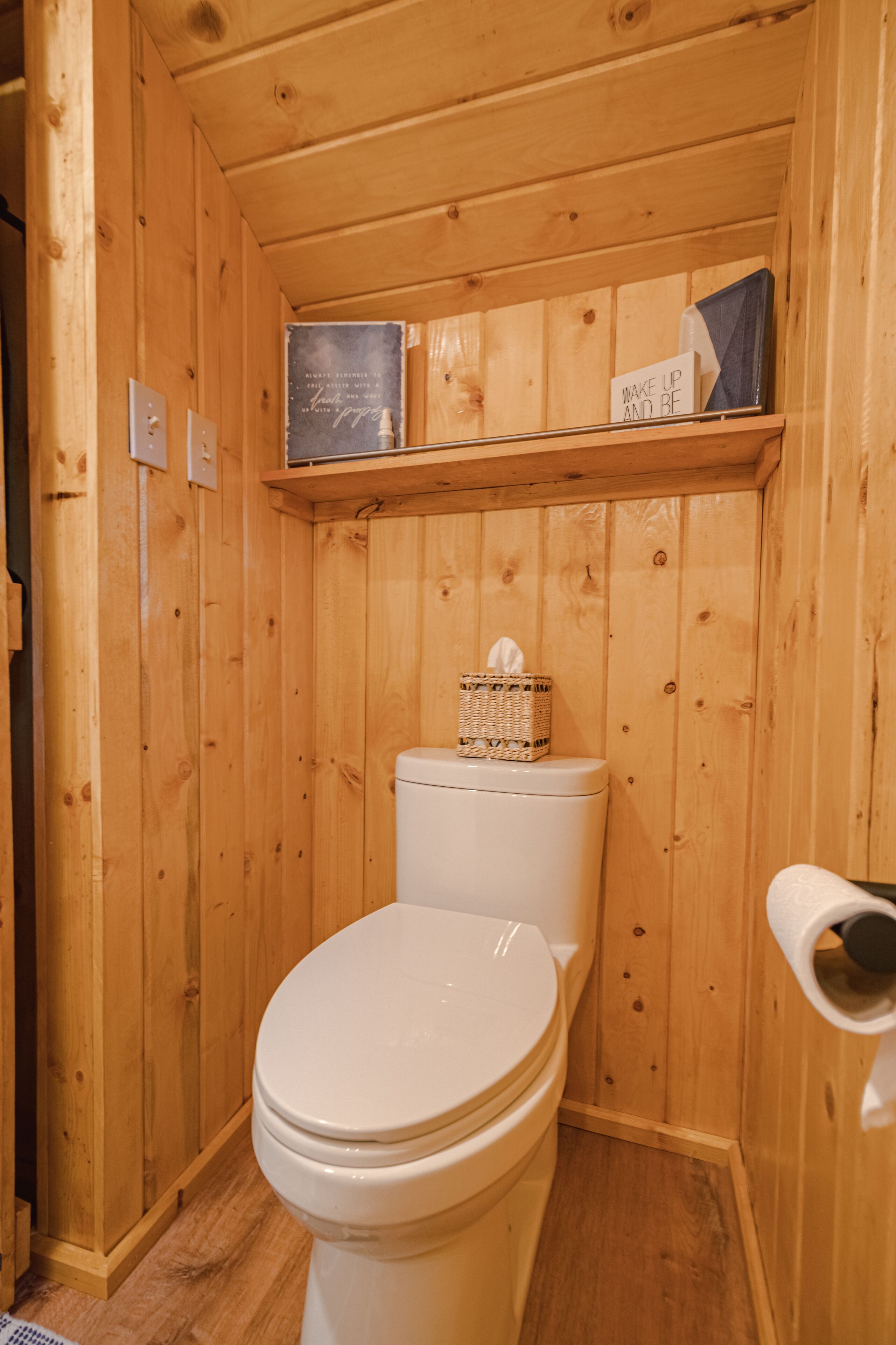 Un baño con paneles de madera, un inodoro, un estante sobre el inodoro y una cesta en el estante.
