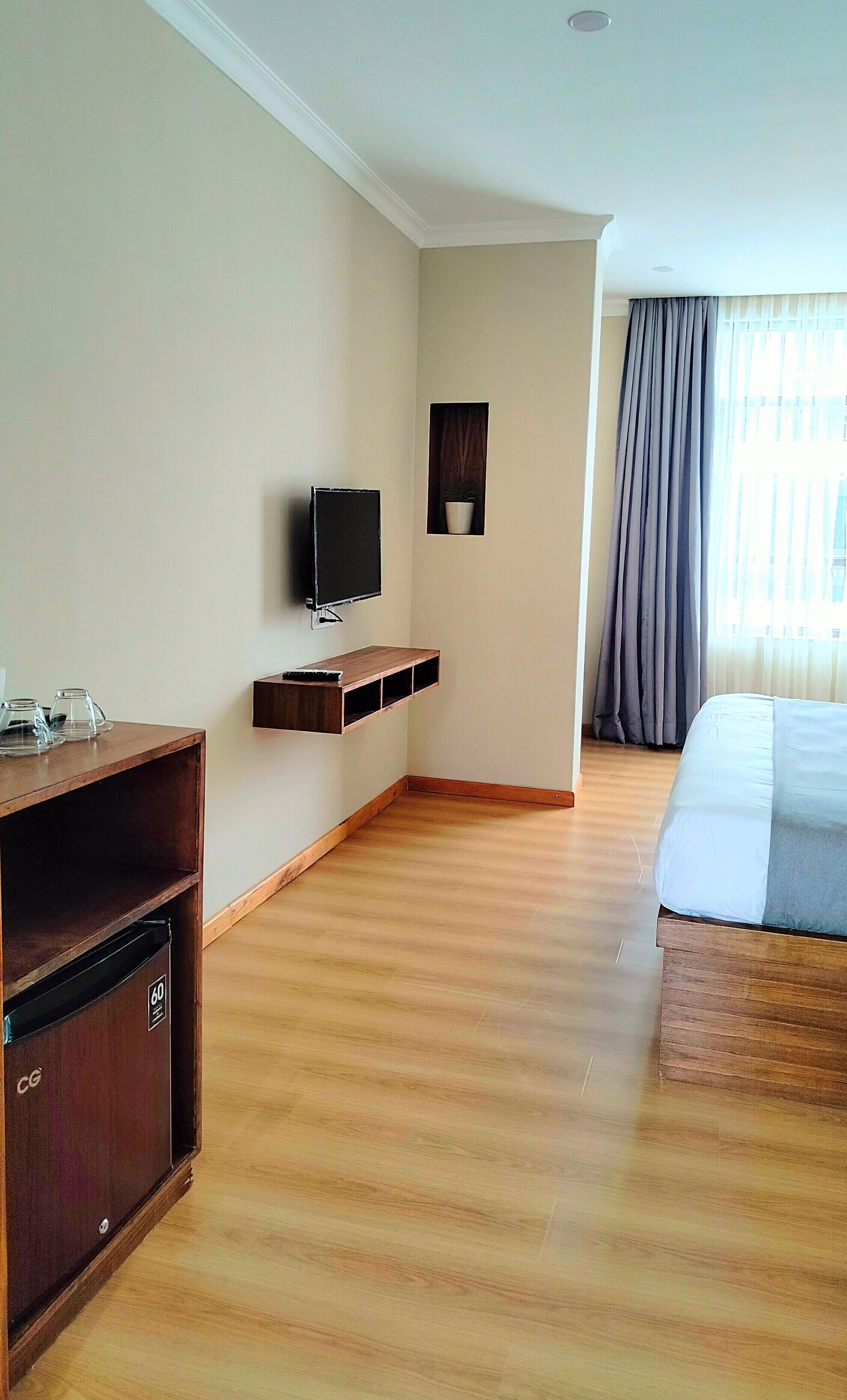 A hotel room with a wooden floor, a bed, a TV mounted on the wall, a small shelf, and a window with curtains.