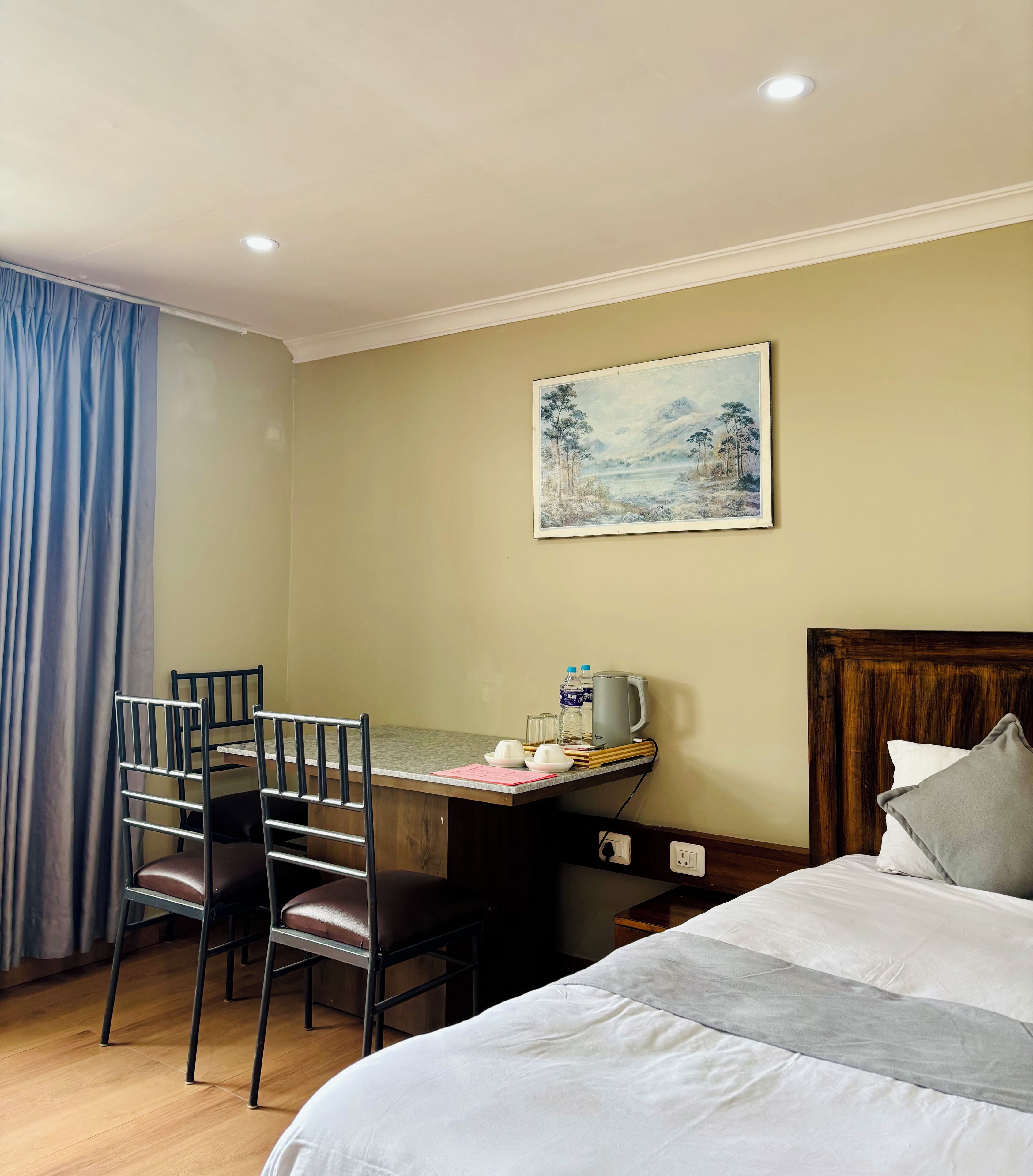 A hotel room with a bed, a desk with a chair, a painting on the wall, and a window with curtains.
