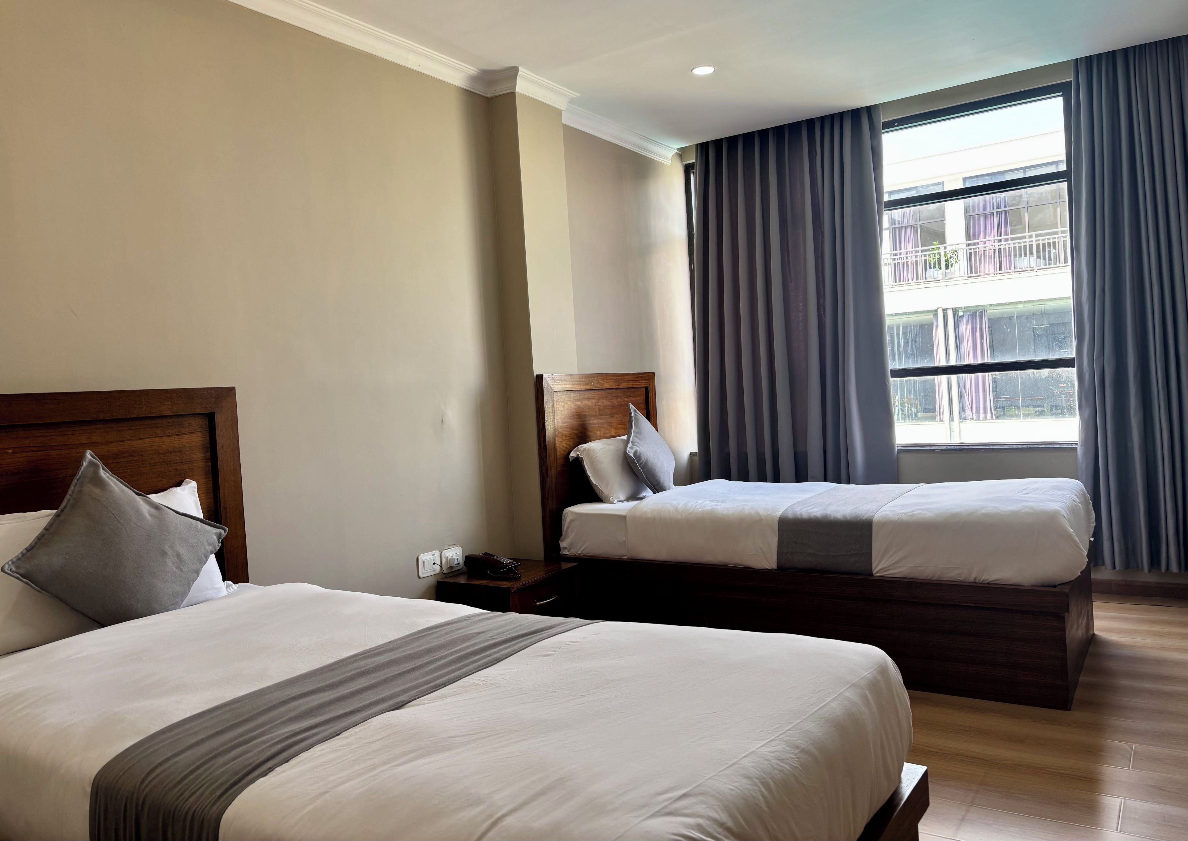 A hotel room with two beds, a nightstand, a window with curtains, and a view of the outside.