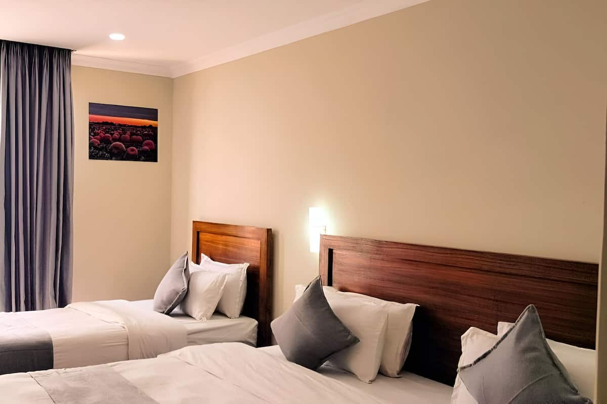 Quarto de hotel com duas camas, uma janela grande com cortinas, um quadro na parede e uma cabeceira de madeira.
