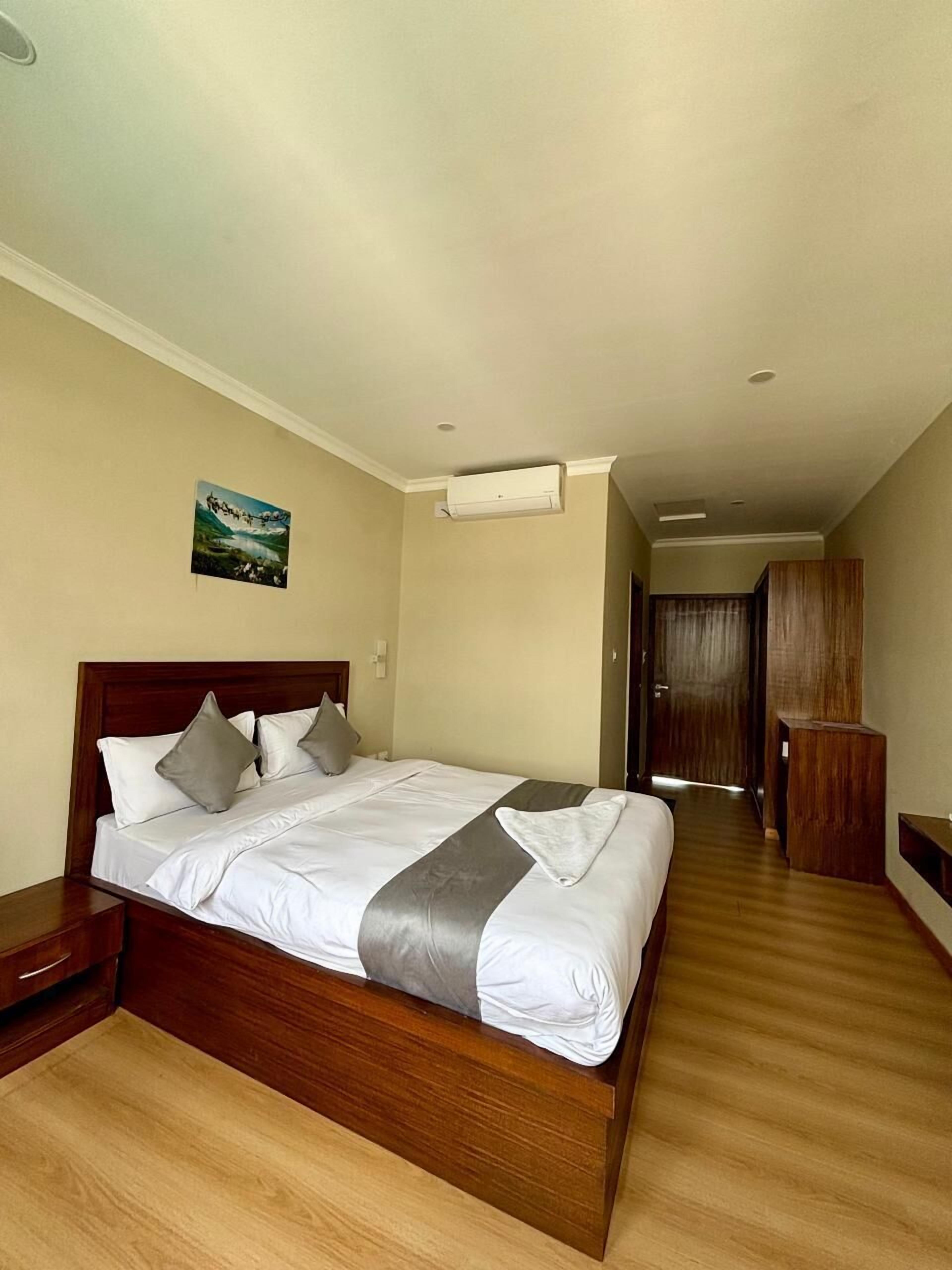A hotel room with a large bed, wooden headboard, a painting on the wall, and a hallway leading to a door.