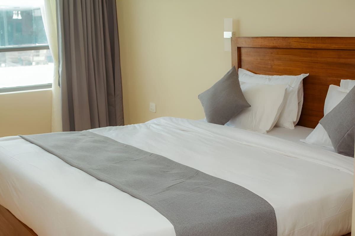 Cama bem arrumada com roupa de cama branca e cinza, cabeceira de madeira e janela com cortinas.