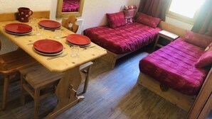Una habitación compacta con una mesa de comedor de madera dispuesta para cuatro personas, dos camas individuales con ropa de cama roja, un pequeño armario de madera y una ventana con cortinas.
