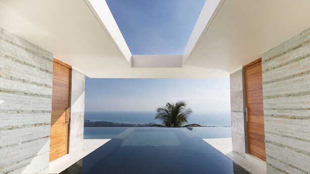A modern infinity pool with a clear view of the ocean and a palm tree, surrounded by white brick walls and wooden doors.