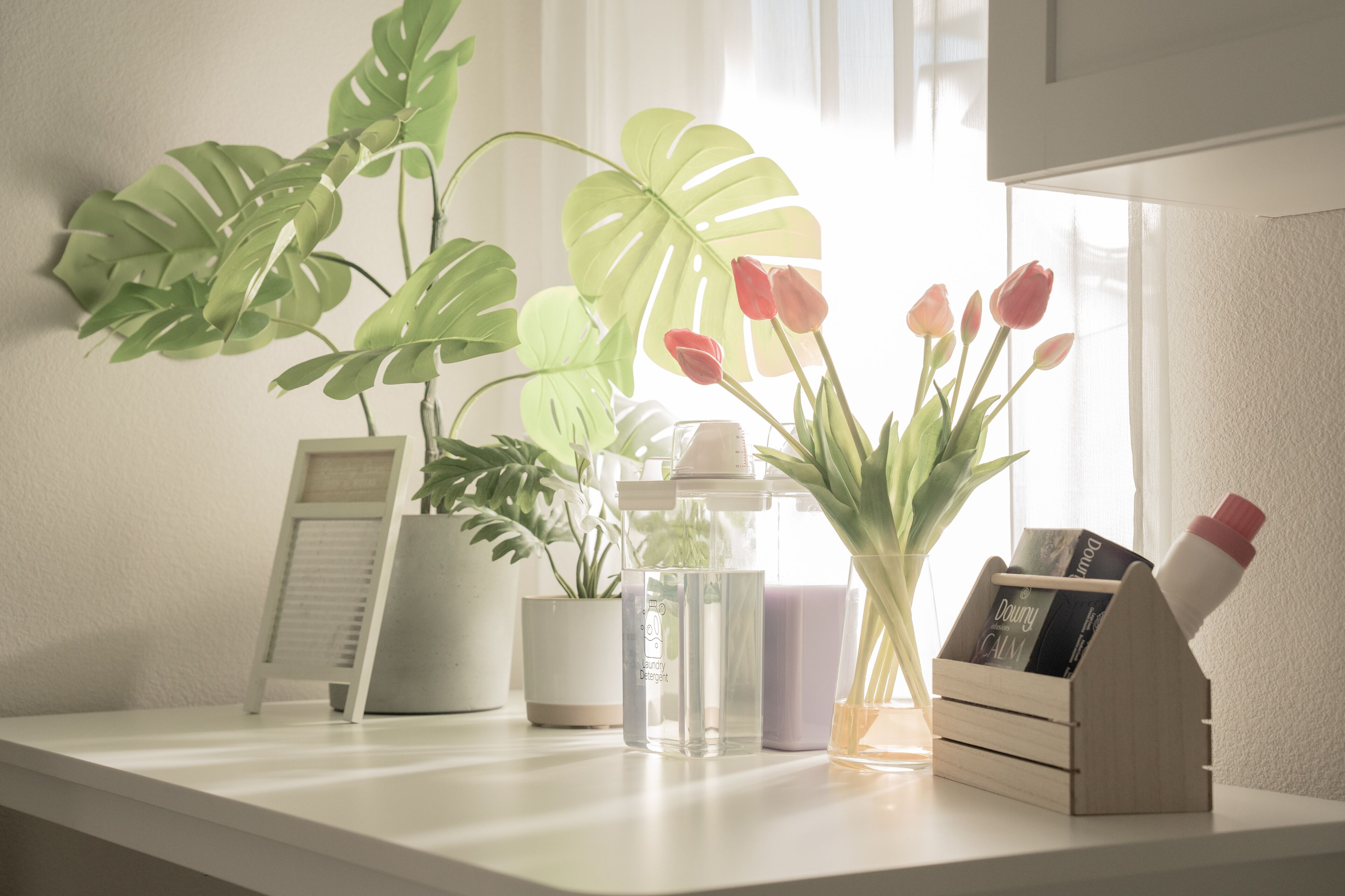 A white countertop with a potted plant, a glass bottle, a small wooden box, and a vase with flowers.