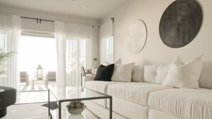 A modern living room with a white sofa, a glass coffee table with a plant, and a view of a balcony with a railing. - Upscale Home - Heated Modern Pool, Outdoor BBQ, Large Kitchen, Up to 8 Guests! (Las Vegas)