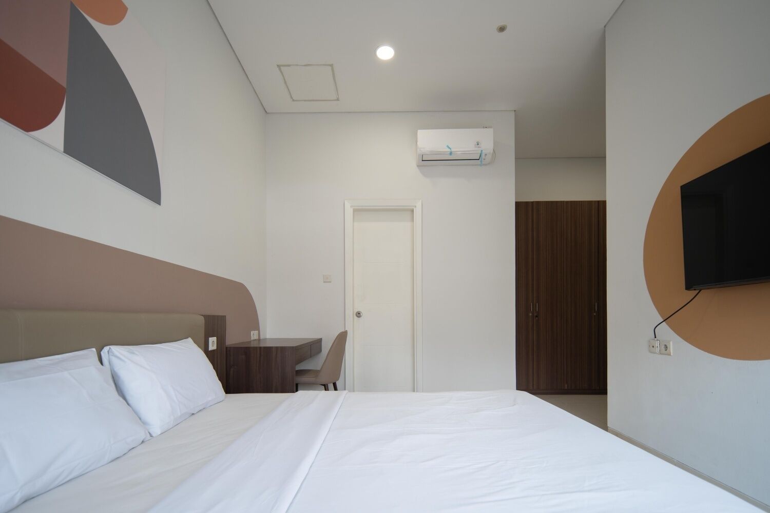 A hotel room with a bed, a desk, a chair, an air conditioning unit, and a television mounted on the wall.