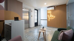 A modern hotel lobby with a reception desk, a green sofa, patterned floor tiles, and a neon art installation. - Cove Senja (Jakarta)