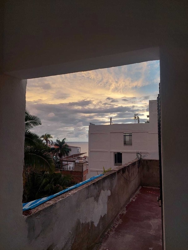 A view through an open doorway to a balcony with a concrete wall, a small window, and a view of the sky with clouds.