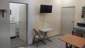 A small, compact room with a tiled floor, a white door, a television mounted on the wall, a small table with a chair, and a microwave on a counter.