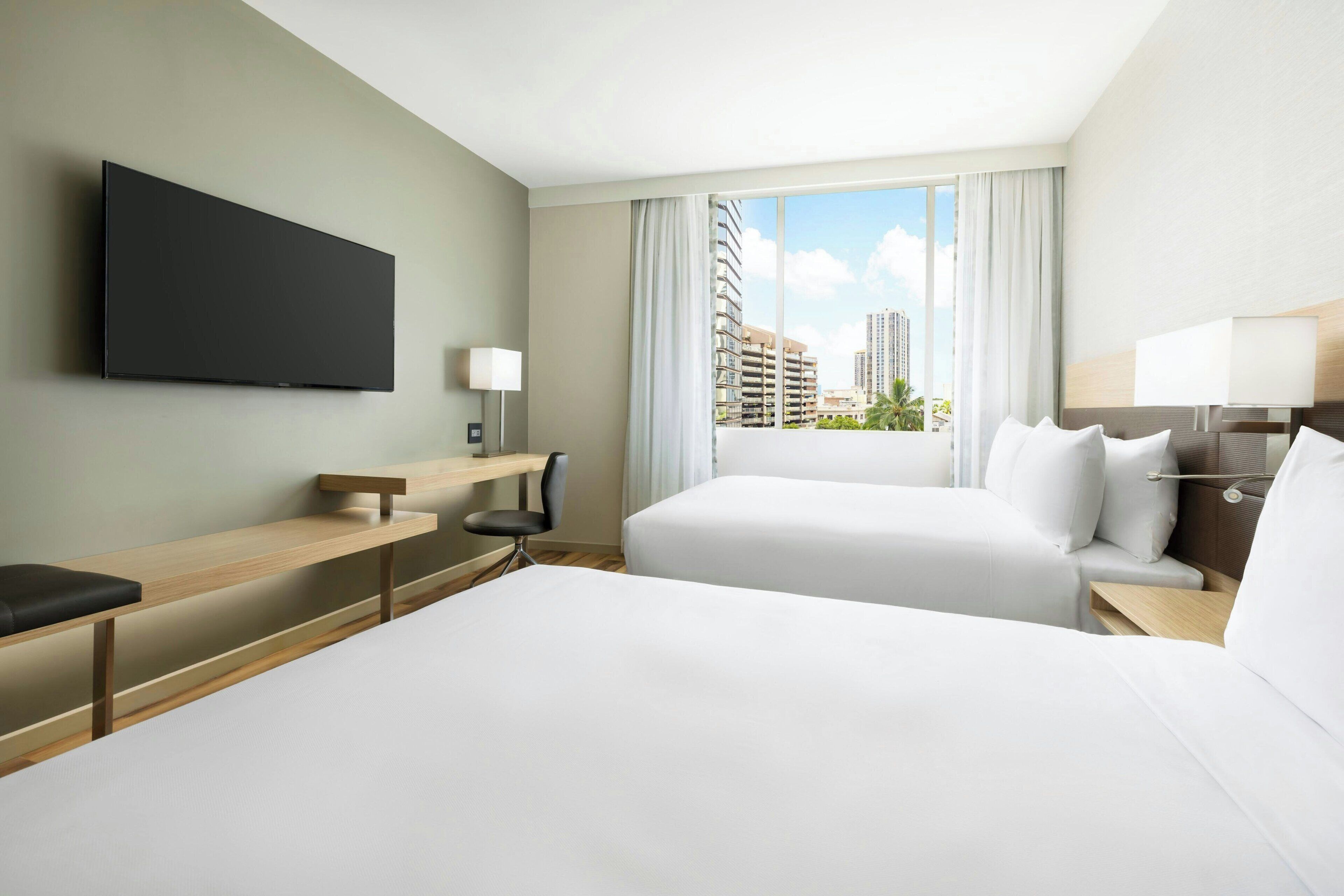 A hotel room with a large bed, a desk, a chair, a TV mounted on the wall, and a window with a city view.