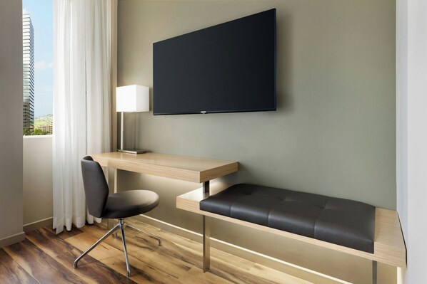 A modern hotel room with a wooden desk, a black chair, a bench, a flat-screen TV, and a window with curtains. - AC Hotel by Marriott Honolulu (Honolulu)