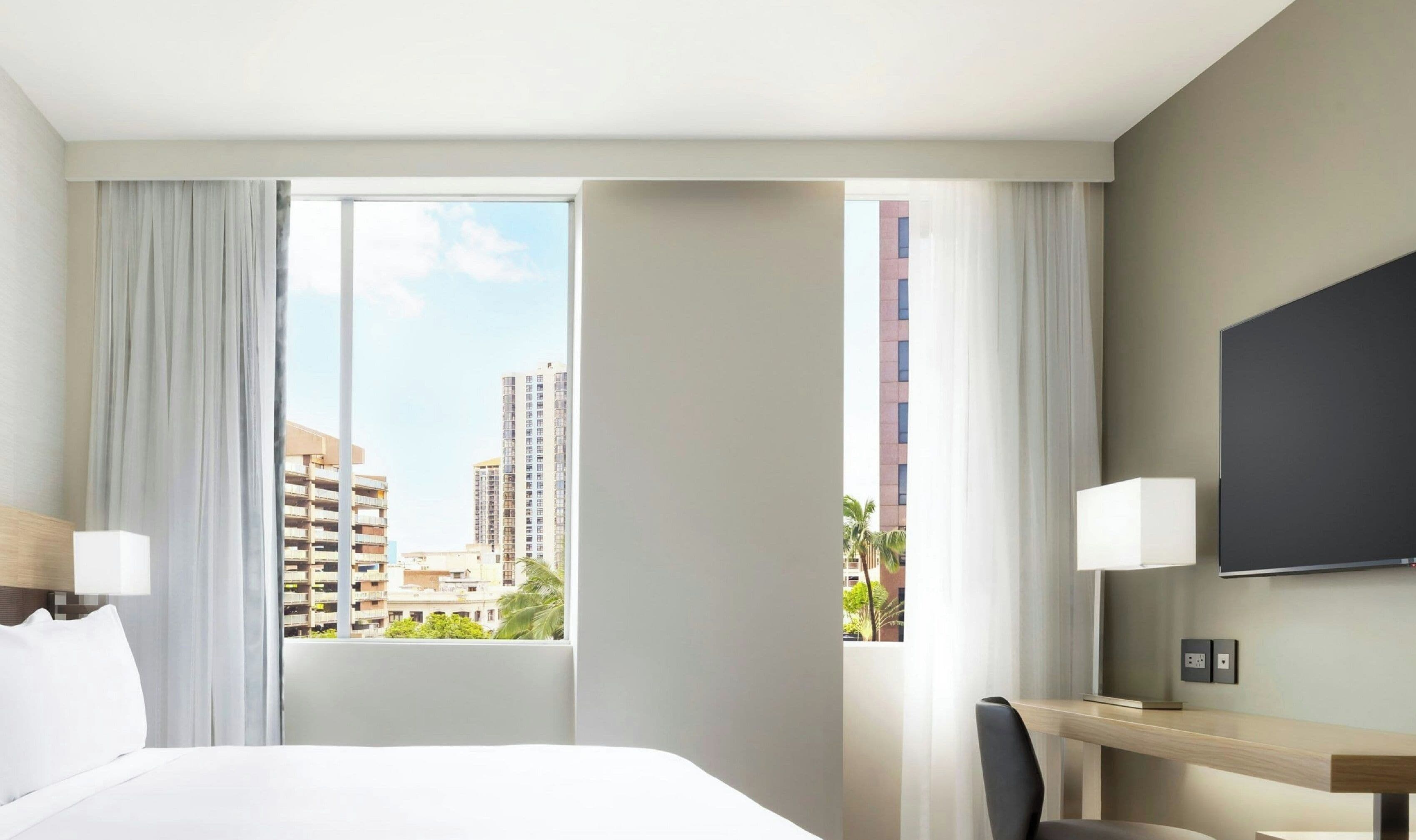 A hotel room with a large bed, a desk with a chair, a TV mounted on the wall, and a window with curtains.