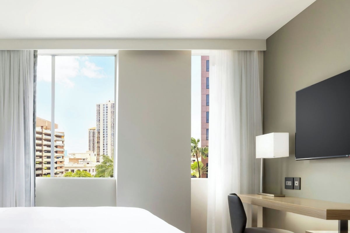 Habitación de hotel con una cama grande, un escritorio con silla, un televisor montado en la pared y una ventana con cortinas.