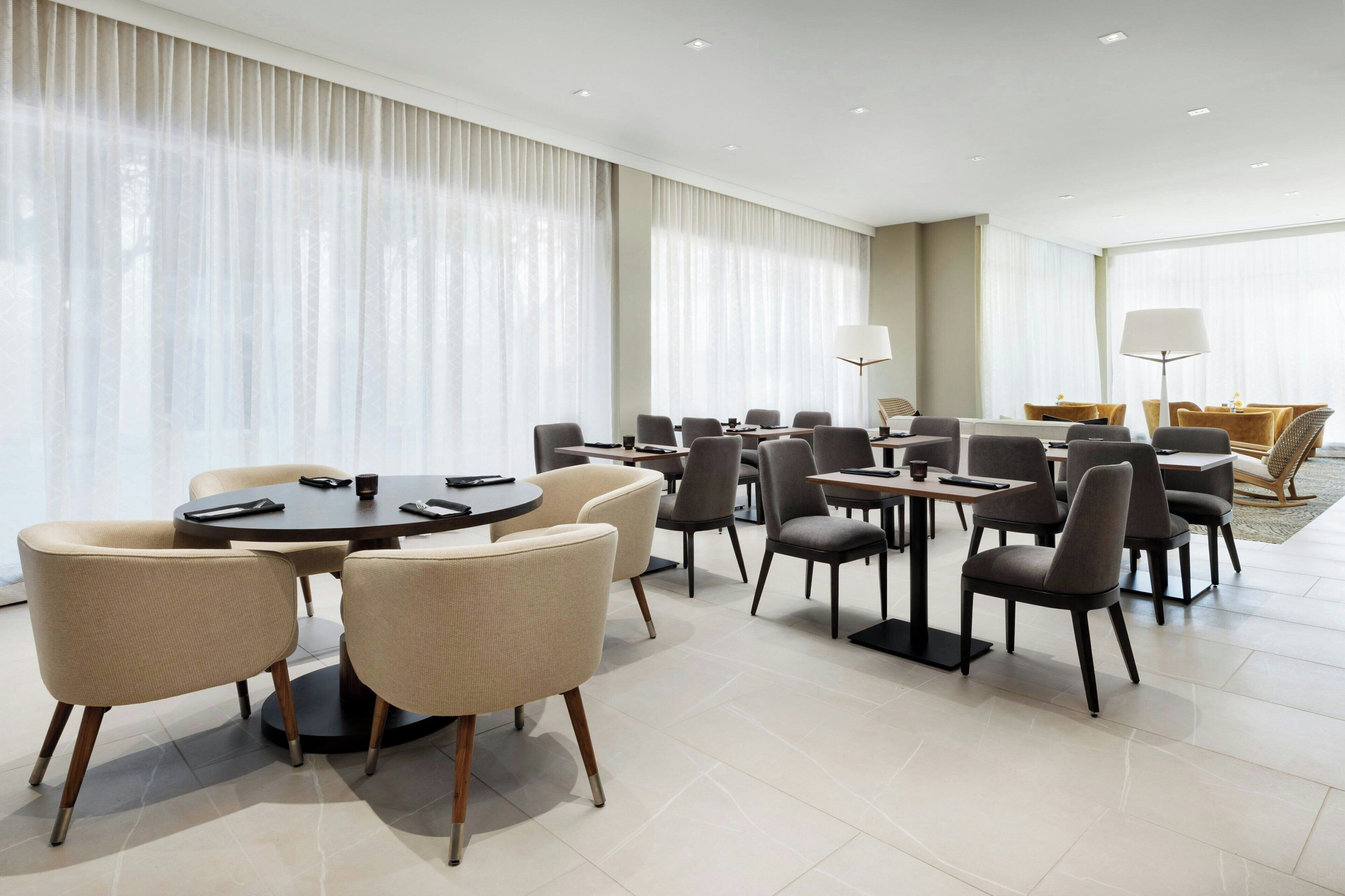 Ein Hotelkonferenzraum mit runden Tischen und Stühlen, großen Fenstern mit transparenten Vorhängen und einer separaten Sitzecke mit Sofas und Lampen.