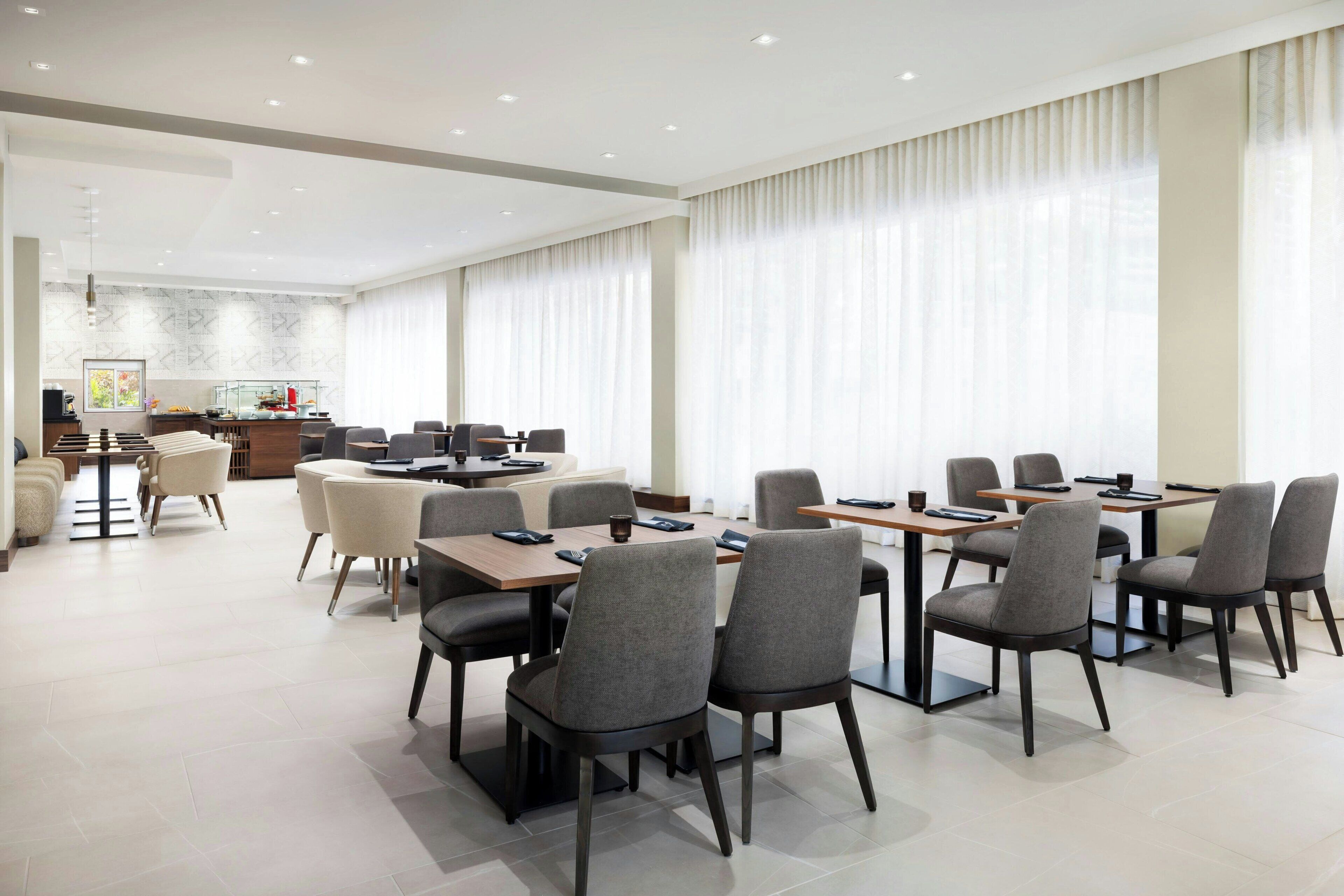 A spacious dining area with multiple tables and chairs, a kitchen in the background, and large windows with curtains.