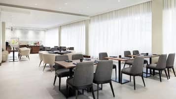 A spacious dining area with multiple tables and chairs, a kitchen in the background, and large windows with curtains.