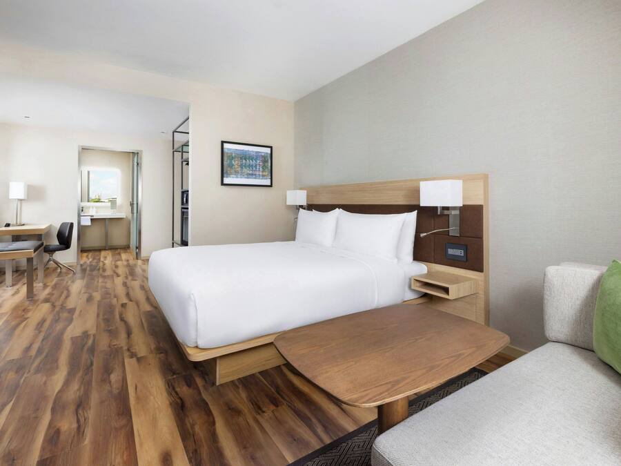 A hotel room with a large bed, a wooden desk, a chair, a small table, and a green pillow.
