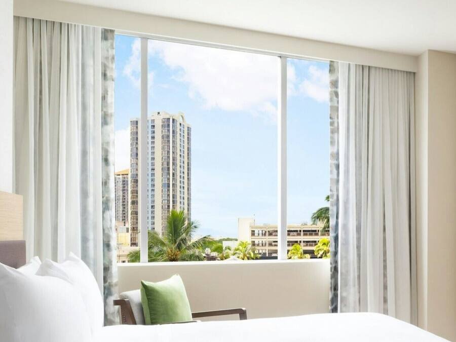 A hotel room with a large window, a bed, and a view of high-rise buildings.