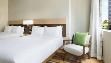 A hotel room with two beds, a wooden headboard, a nightstand with a lamp, a chair with a green pillow, and a window with curtains.