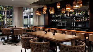 A restaurant with a long wooden table and wicker chairs, a bar area with shelves of bottles, and hanging wicker lamps. - AC Hotel by Marriott Honolulu (Honolulu)