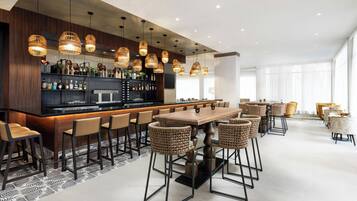 Eine moderne Bar mit einer langen Theke, mehreren Pendelleuchten und verschiedenen Sitzgelegenheiten, darunter Barhocker und hohe Stühle.