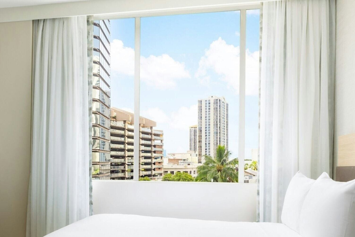A hotel room with a large bed, white curtains, and a view of tall buildings and palm trees.
