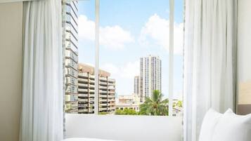 A hotel room with a large bed, white curtains, and a view of tall buildings and palm trees.