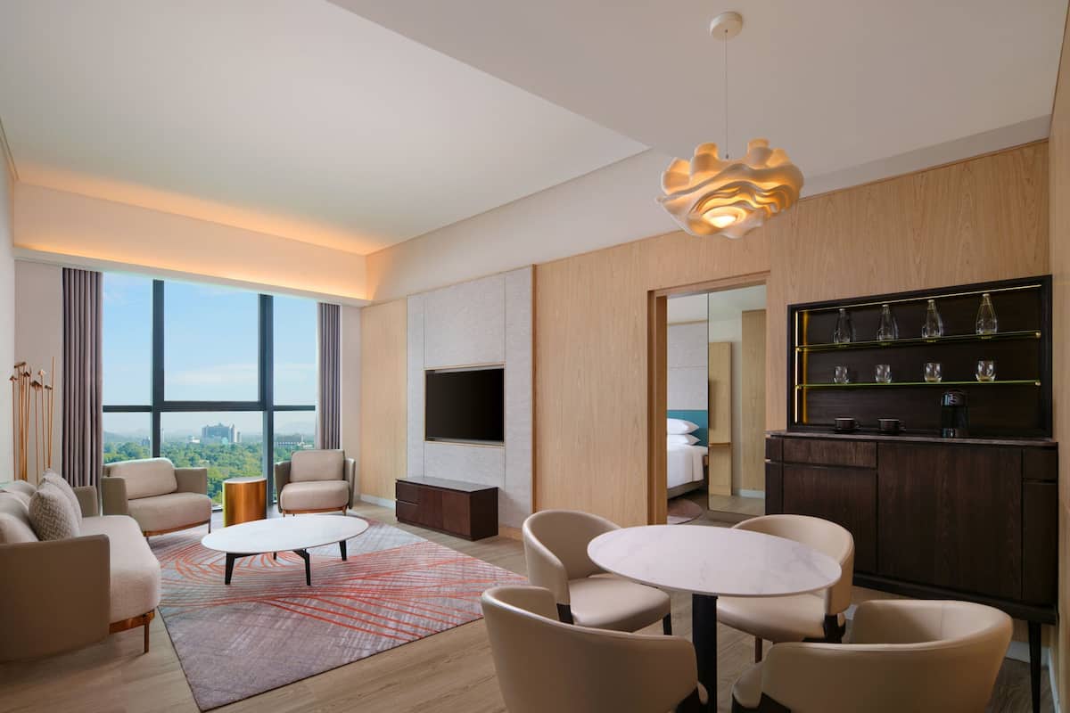 A modern hotel room with a large window, a flat-screen TV, a dining area, and a minibar.