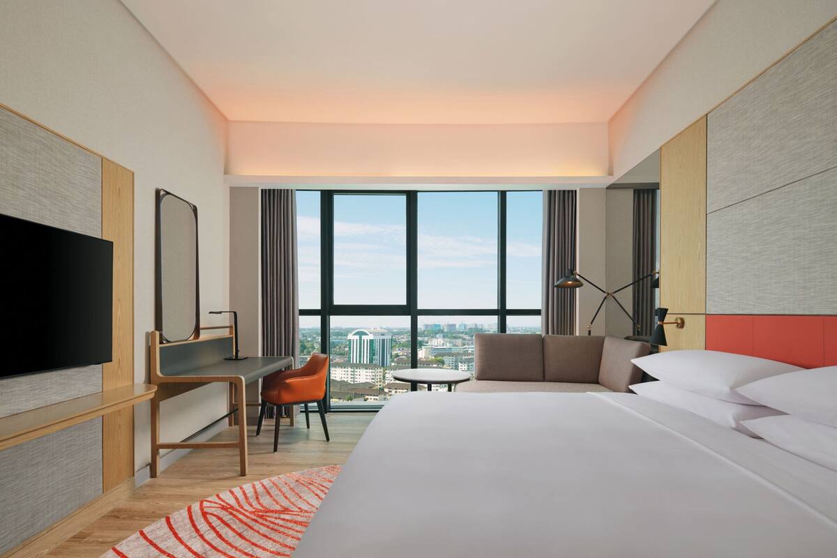 A modern hotel room with a large bed, a desk, a chair, a TV, and a view of the city through large windows.