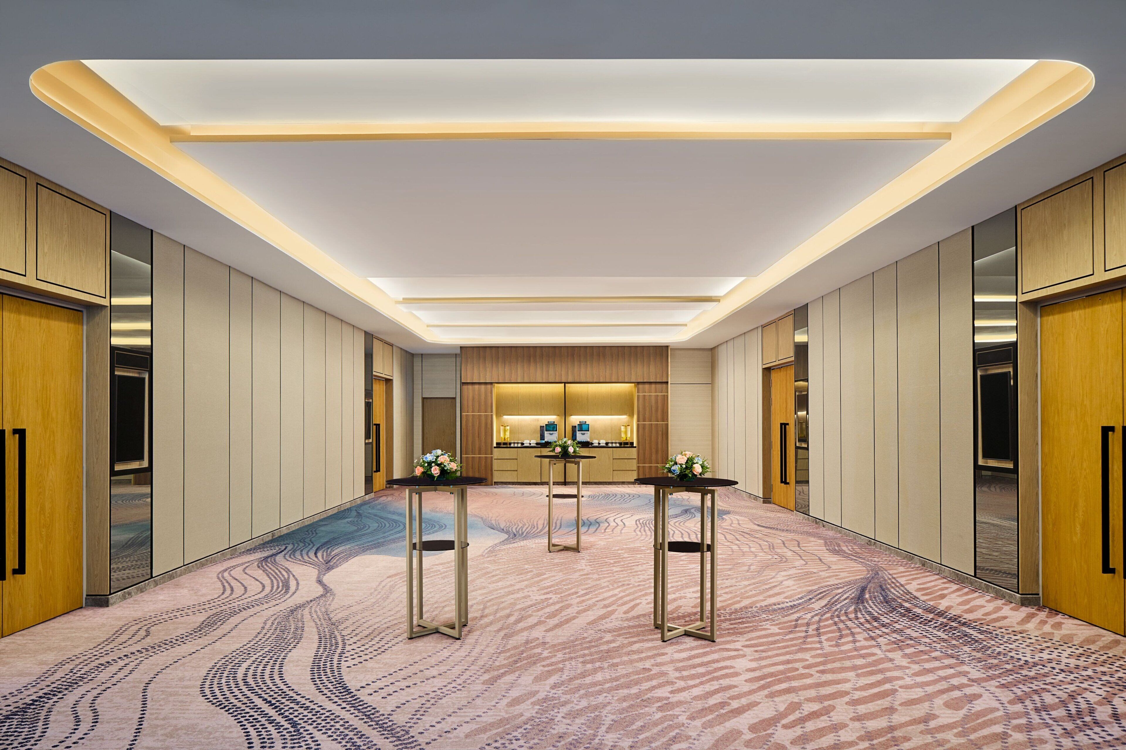 Un couloir d’hôtel moderne, avec un tapis à motifs, des casiers en bois et un espace de réception comprenant un comptoir et des chaises.