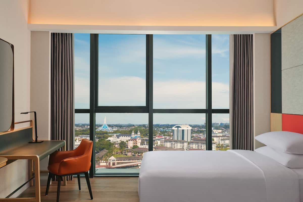 A hotel room with a large window, a bed, a desk, and an orange chair.
