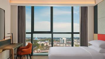 A hotel room with a large window, a bed, a desk, and an orange chair.