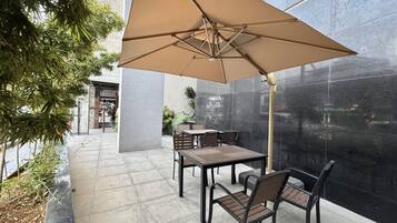 An outdoor patio area with a table and chairs under a large umbrella, surrounded by a stone wall and greenery.