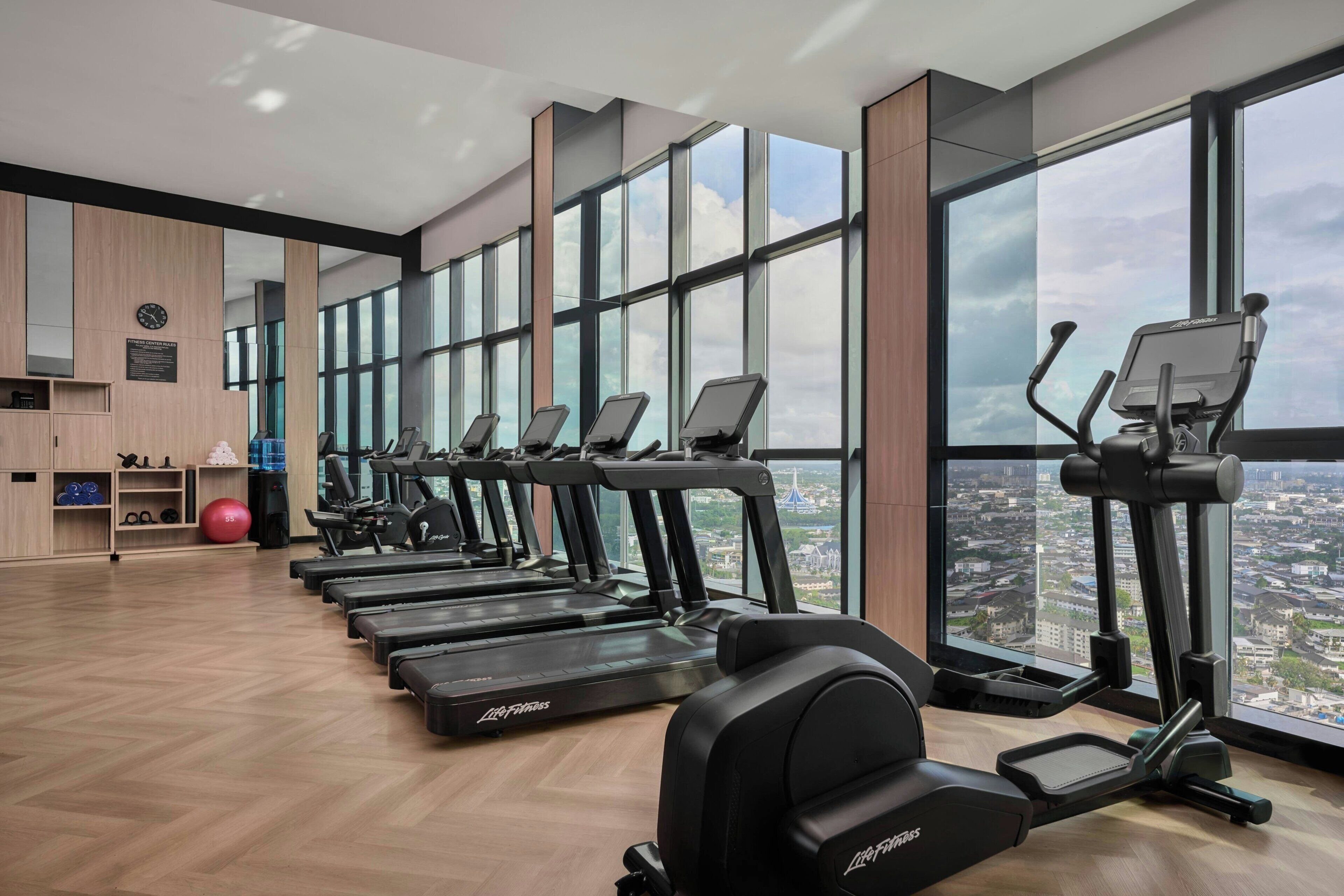 Un gimnasio con varias cintas de correr y equipo de ejercicio, ventanas amplias y vista panorámica de la ciudad.
