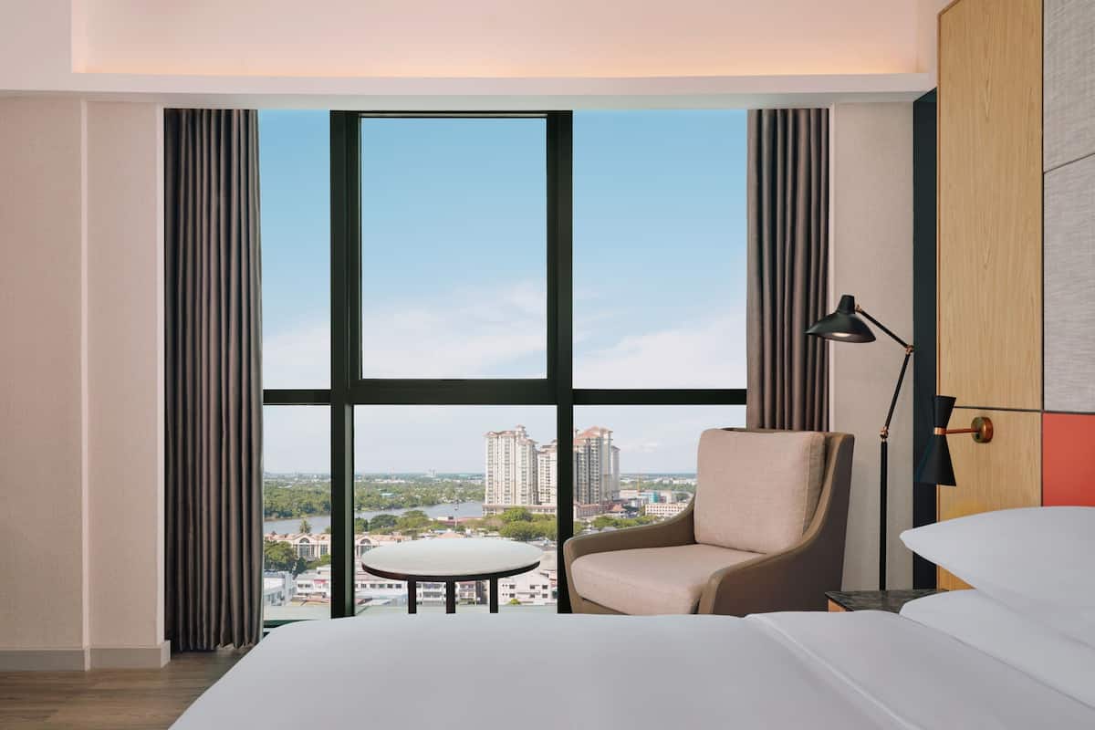 A hotel room with a large window, a bed, a chair, a small table, and a lamp.